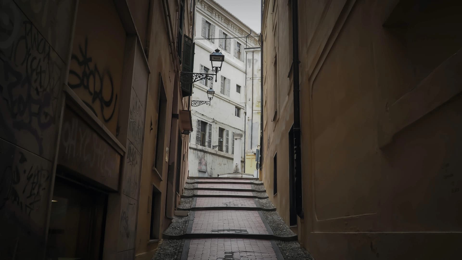 A dark, narrow, inclined path encompassed by a row of buildings on either side leading to the light of day by Raphael Biscaldi